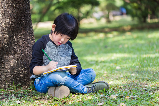 Little Asian Boy Writing Book With Pencil In The Park