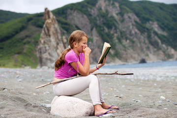 Girl reading book sitting on a stone on a sandy sea shore