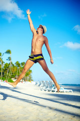 Happy muscled man in sunhat on beach jumping behind sky