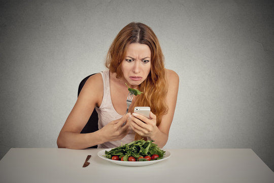 Displeased girl eating green salad looking at phone bad news