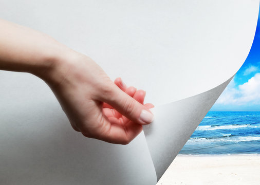 Hand pulling a paper corner to uncover, reveal sunny beach