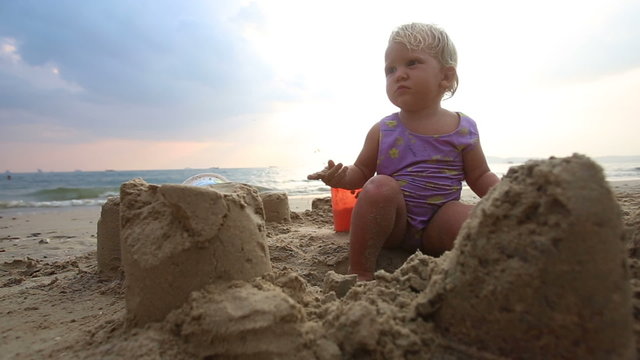 small baby girl in purple swimsuit play in the sand castle on th