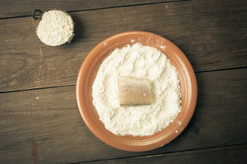 One piece of fresh fish with flour on a plate on a wooden backgr