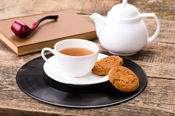 ivory tea cup with sweet cookie, book and tobacco pipe on wooden