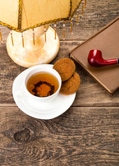 ivory tea cup with sweet cookie, book and tobacco pipe on wooden