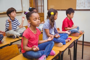 Mixed race pupils meditating in lotus position on desk in classroom - obrazy, fototapety, plakaty