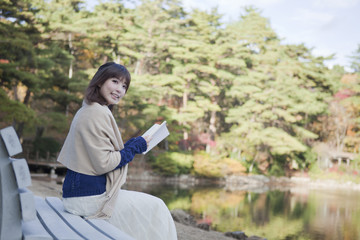 秋の紅葉の公園の池の前のベンチで本を読む女性