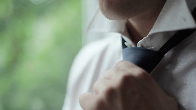Man in white shirt tying a tie near the window. Close-up