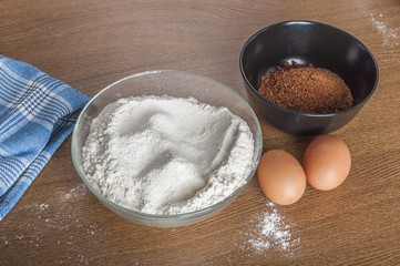 Flour in a bowl, eggs and brown sugar on wooden background - 77023785