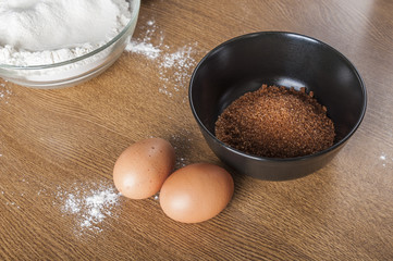 Flour in a bowl, eggs and brown sugar on wooden background