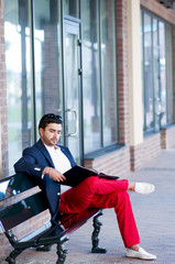 Stylish young man resting on a bench, reading  a magazine.