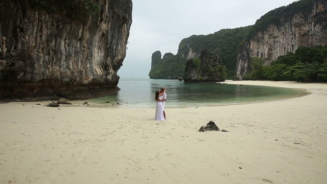 blonde bride and young groom kiss standing on white sand beach 