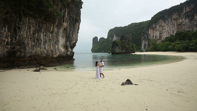 blonde bride and young groom kiss standing on white sand beach 