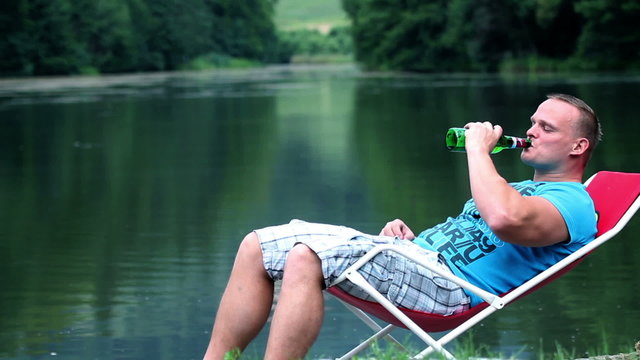 Young man lying in a deckchair near lake and drinking refershing bevarage