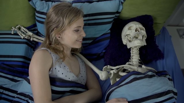 Young woman wakes up in bed next to a skeleton and hugs him 