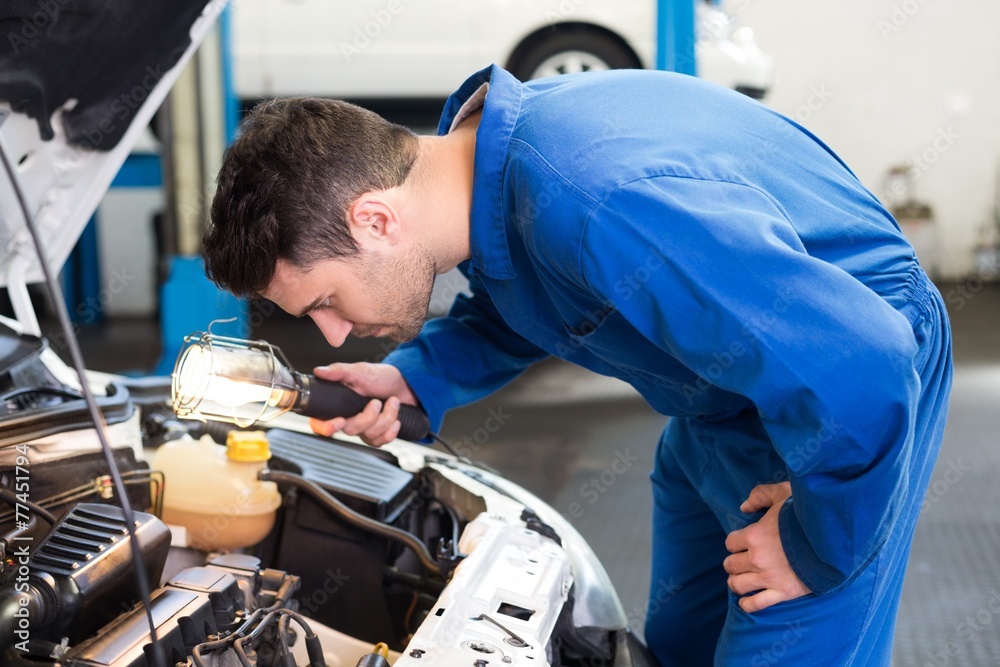 Poster mechanic examining under hood of car with torch