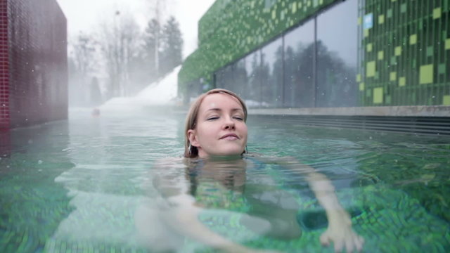 Shot of a pretty young woman in blue bikini that swimms towards the camera, enoying in the outdoor pool when snowing