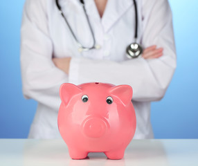 Doctor with pink piggy bank on blue background, close up
