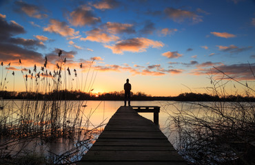 Man standing on a jetty during a winter sunset over a lake.
