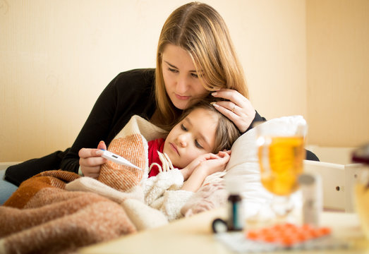 mother checking temperature of sick daughter lying in bed