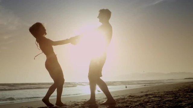 couple holding hands spinning on beach - silhouette of man and woman dancing by ocean on sand in slow motion 4K