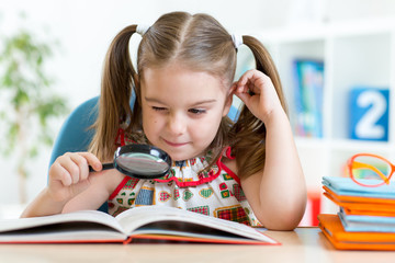 funny child reads book using magnifier sitting at table