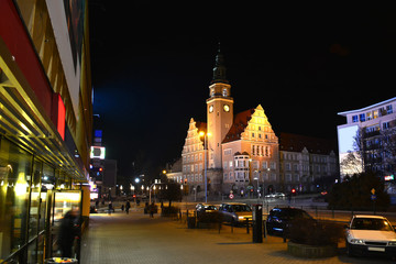 Obraz premium A striking night view of the New Town Hall in Olsztyn, Poland. The illuminated Neo-Gothic building with its tall clock tower dominates the city center under a dark evening sky.