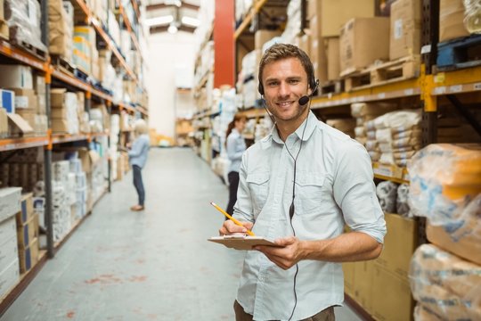 Warehouse manager wearing headset writing on clipboard - Powered by Adobe