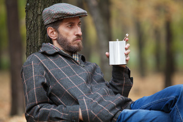 Man with a beard sitting in the autumn forest with a flask in hi