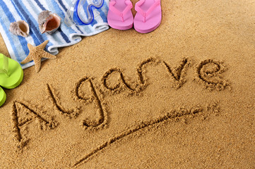 The word Algarve written in sand on a beach with towel flip flops seashells and starfish Portugal...