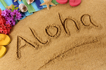 The word Aloha written in sand on a beach with towel flip flops seashells Hawaii summer vacation holiday photo - obrazy, fototapety, plakaty