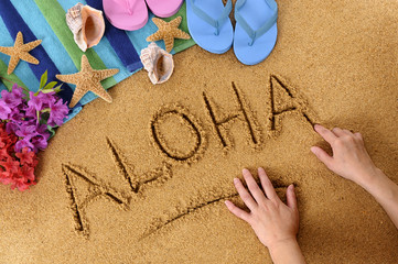 The word Aloha being written in sand by child hands on a beach with towel flip flops seashells...