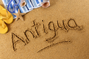 Fototapeta na wymiar The word Antigua written in sand on a beach with towel flip flops seashells summer vacation holiday photo