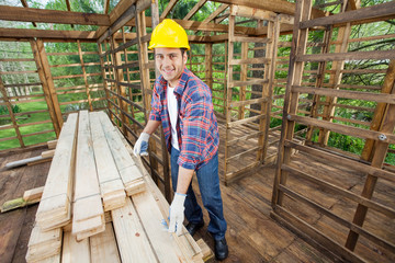Naklejka na ściany i meble Smiling Carpenter Measuring Wooden Plank At Construction Site