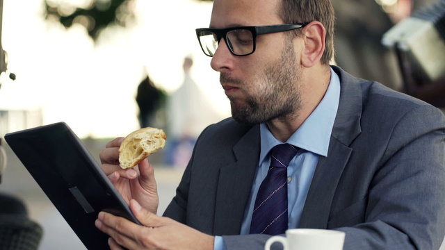 Young businessman with tablet computer eating croissant in cafe