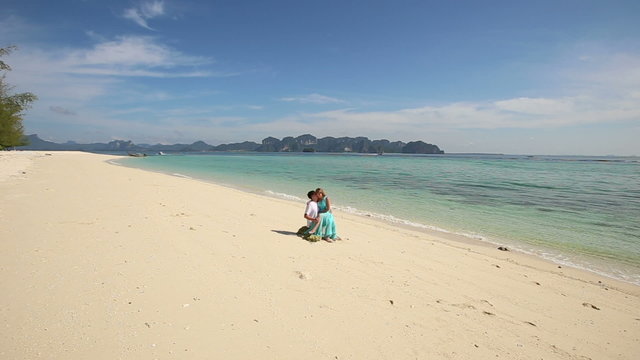 guy sits on stone with asian girl on his knees on sand beach at 