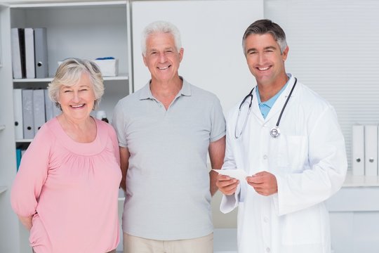 Happy Senior Couple With Doctor Standing In Clinic