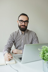 Smiling man with beard in glasses taking notes with laptop notep