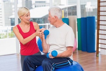 Female trainer assisting senior man in lifting dumbbells