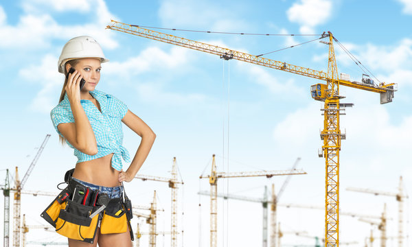Woman in helmet and tool belt, talking on phone. Tower cranes as