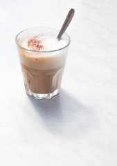 Foamy cappuchino in coffee glass with spoon on white