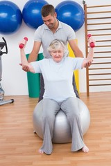 Fototapeta na wymiar Trainer helping senior woman lift dumbbells on exercise ball
