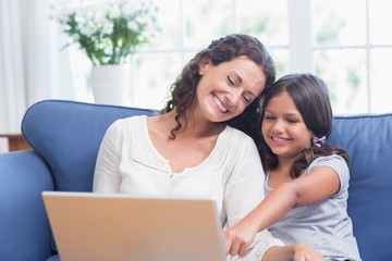 Fototapeta premium Happy mother and daughter sitting on the couch and using laptop