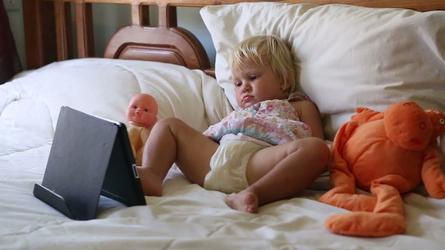 little girl lies in bed among toys and watches cartoons	