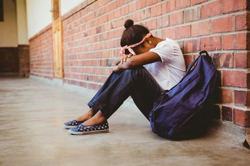 Tensed mixed race girl sitting against brick wall in school corridor