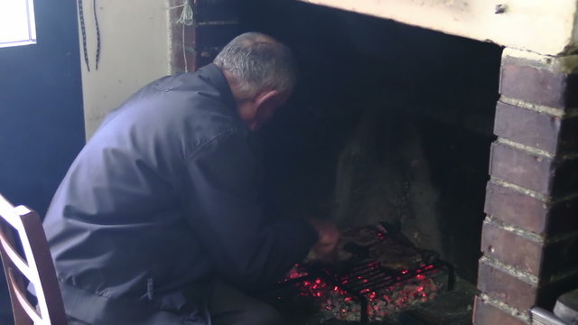 old man cooks steaks on the grill in front of a fireplace