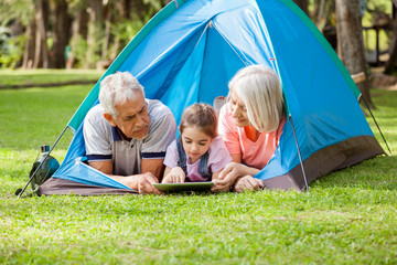 Grandparents With Granddaughter Using Digital Tablet At Campsite