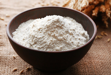 Fototapeta na wymiar Flour in bowl with ears and grains on sackcloth background