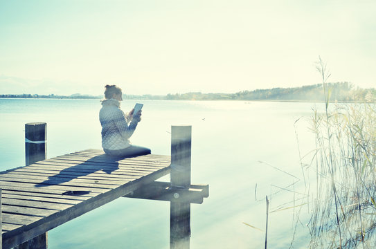 Girl reading from a tablet on the wooden jetty against a lake. S