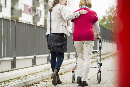 Adult granddaughter assisting her grandmother walking with wheeled walker, back view